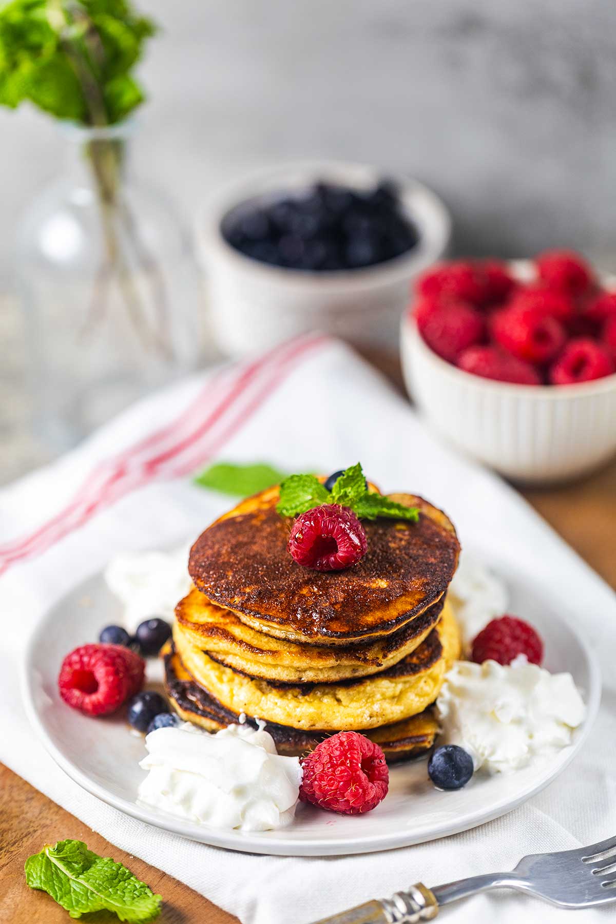 A stack of pancakes topped with a raspberry and mint sits on a white plate, surrounded by whipped cream, raspberries, and blueberries. Bowls of berries and a vase with mint are in the background.