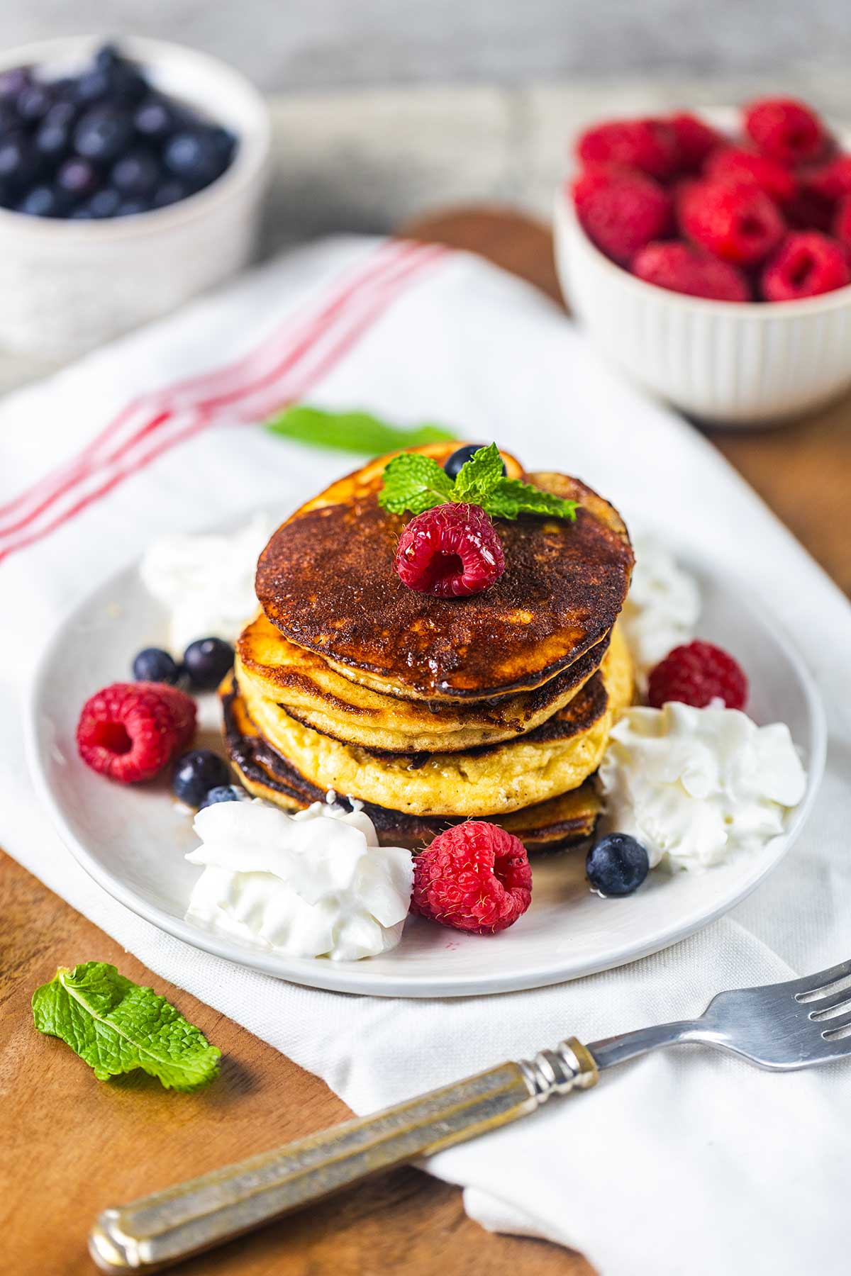 A plate of pancakes topped with raspberries, blueberries, and mint, surrounded by whipped cream, with bowls of blueberries and raspberries in the background. A fork rests on the plate.