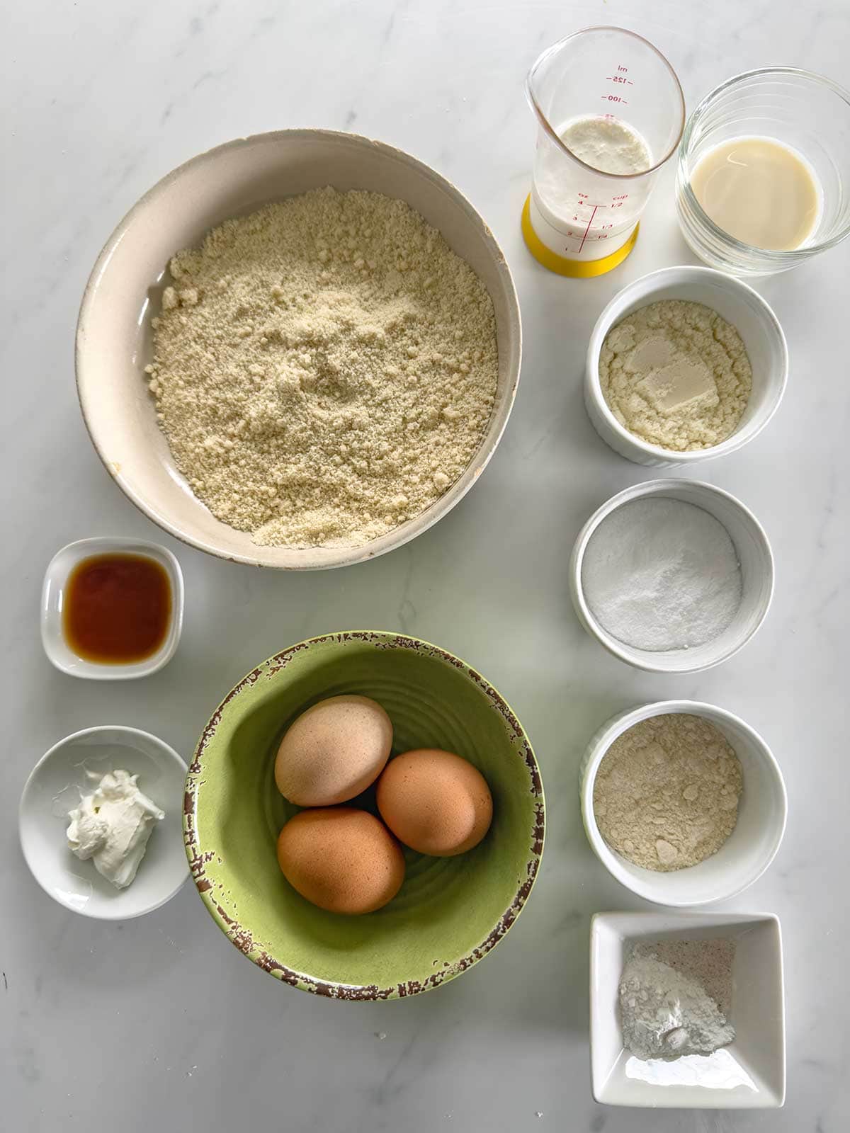 Various baking ingredients are arranged on a white surface, including three eggs in a green bowl, a large bowl of almond flour, small bowls of different powders and liquids, and a measuring cup with a small amount of liquid.