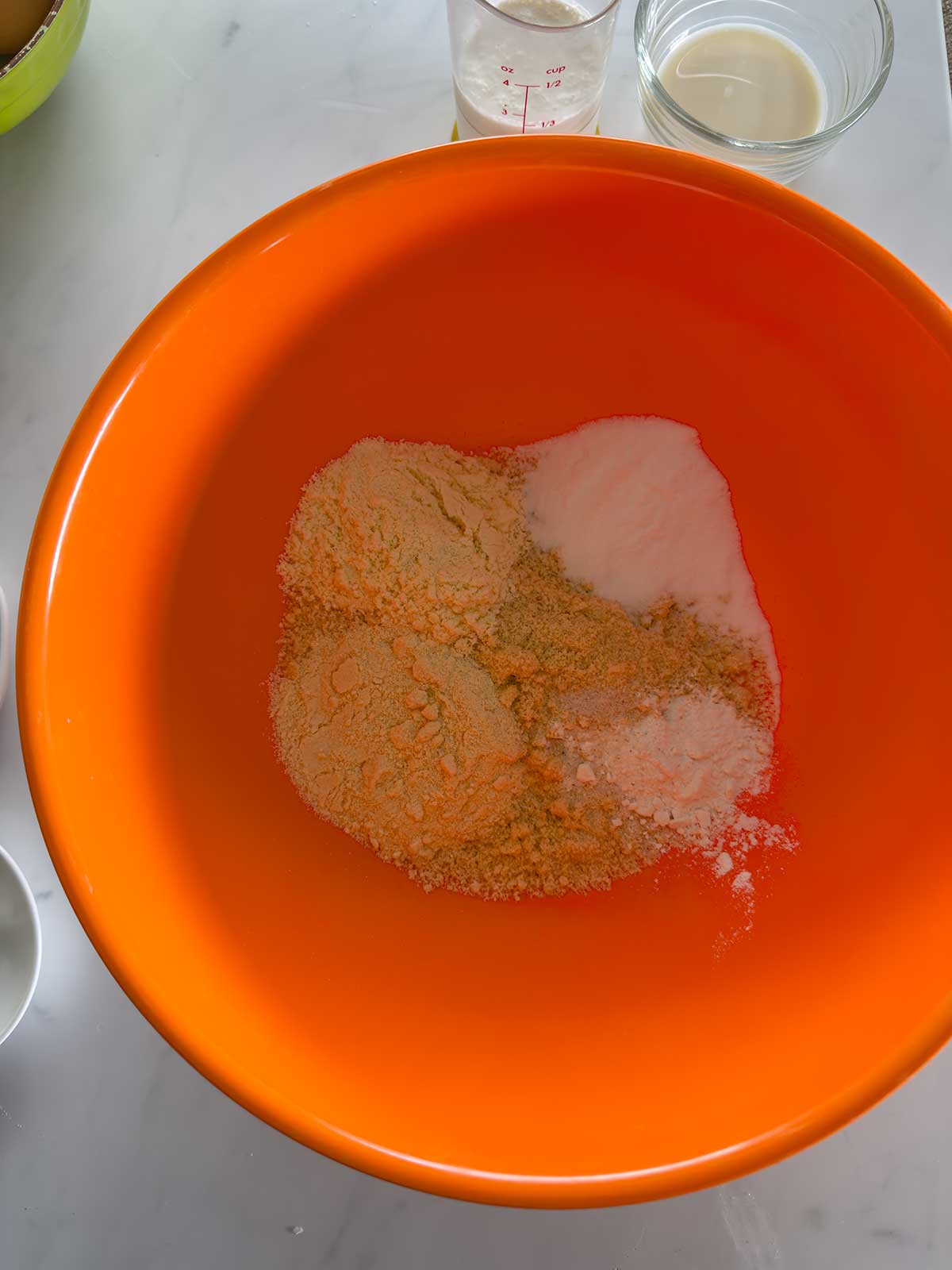 An orange mixing bowl containing dry baking ingredients sits on a white countertop. Small glass bowls with other ingredients are nearby.