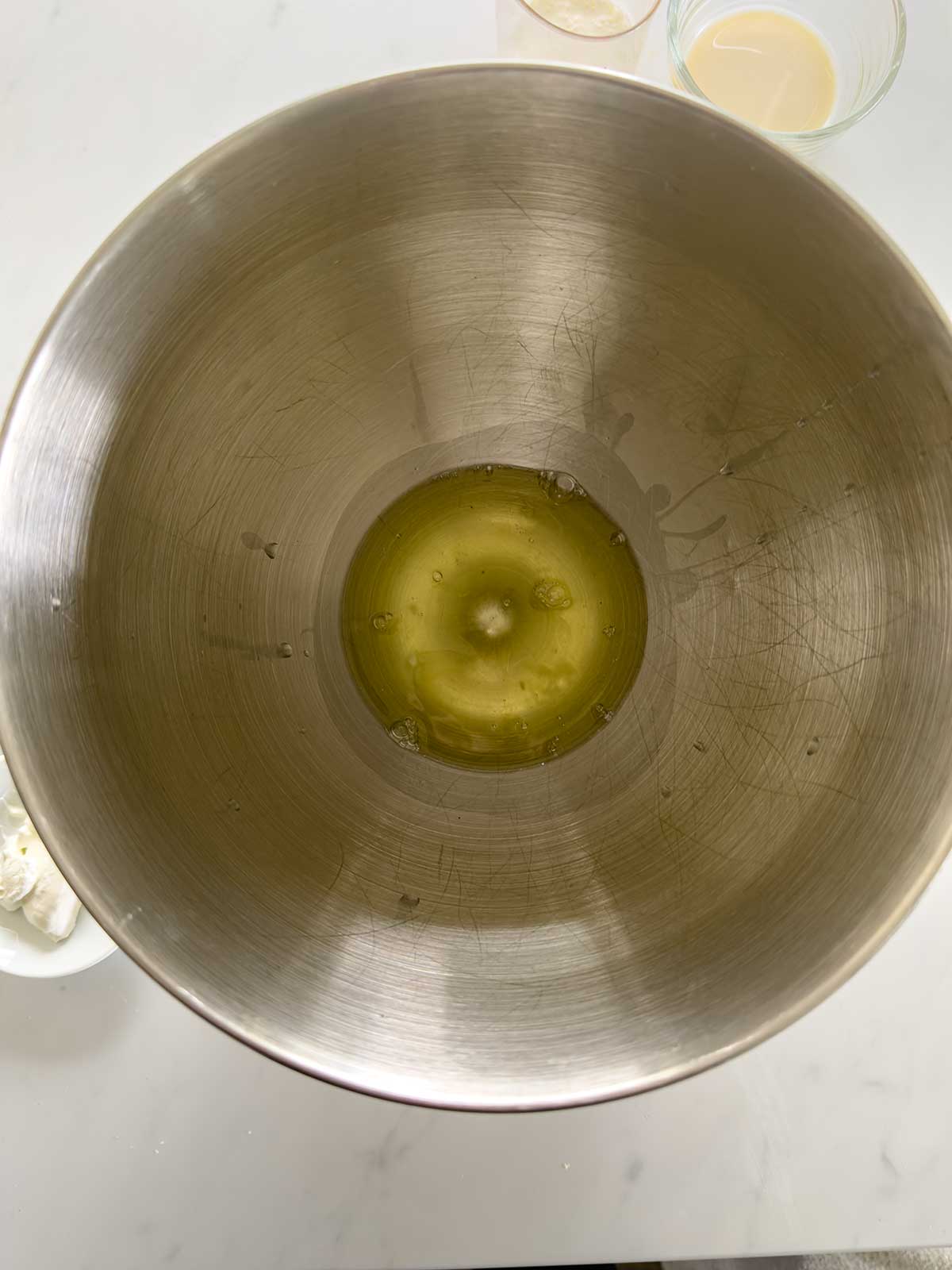 A single raw egg white sits at the bottom of a large stainless steel mixing bowl on a white countertop, with small bowls of ingredients nearby.