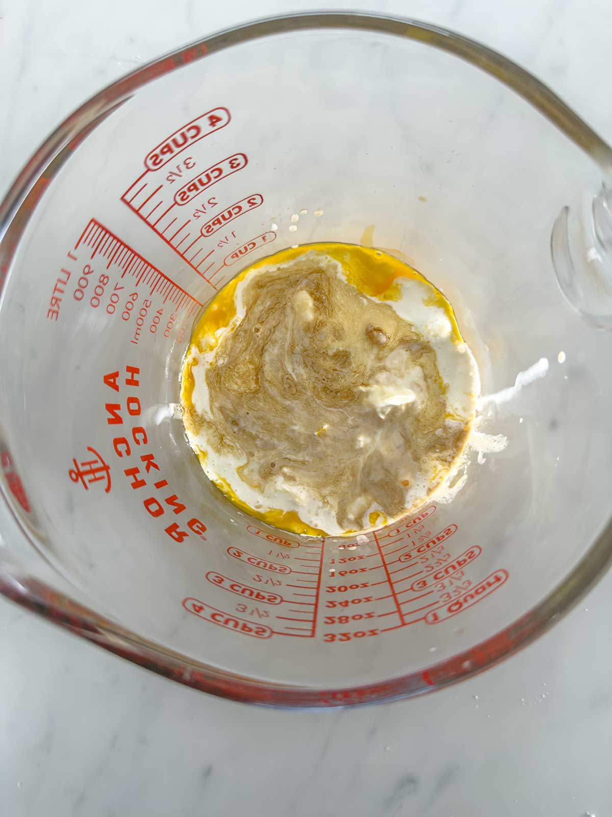 A glass measuring cup on a marble surface contains a mixture of milk, egg yolk, and brown spices, partially blended together. Red measurement markings are visible on the cup.