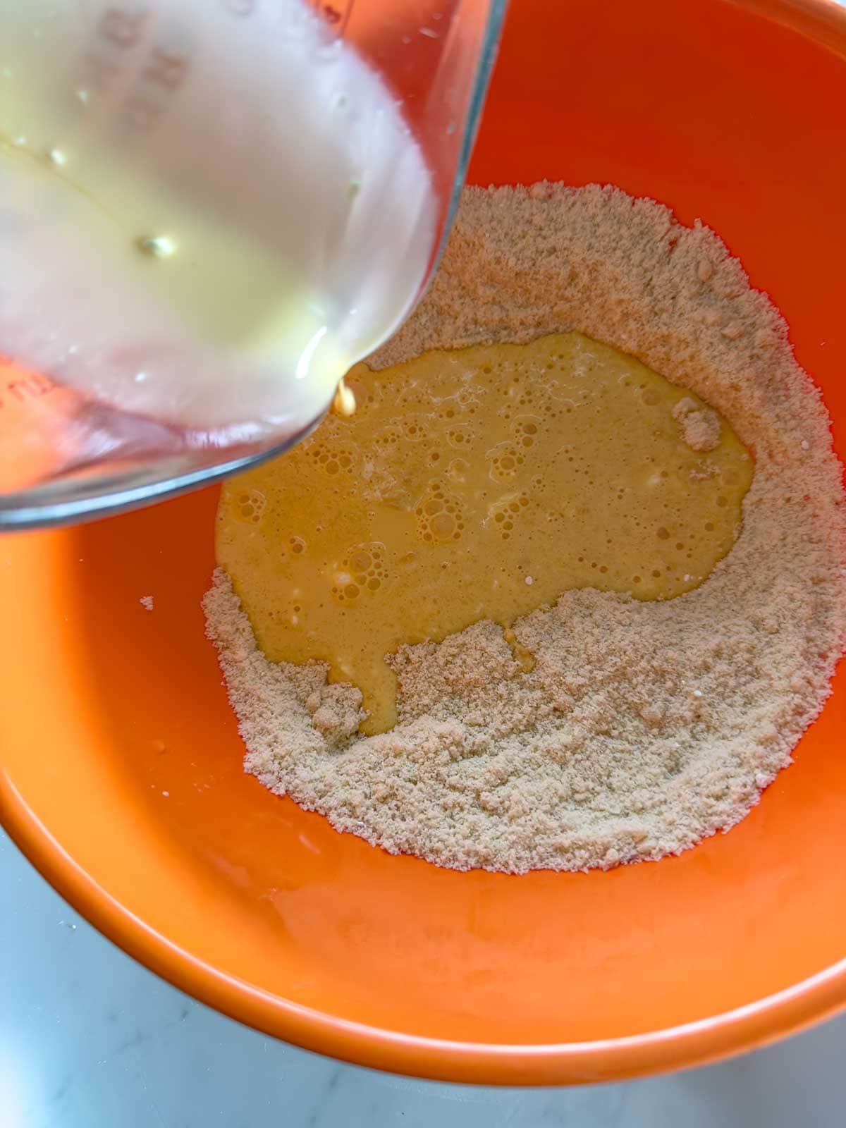A close-up of milk and eggs being poured from a measuring cup into a bowl of dry ingredients, all in an orange mixing bowl.