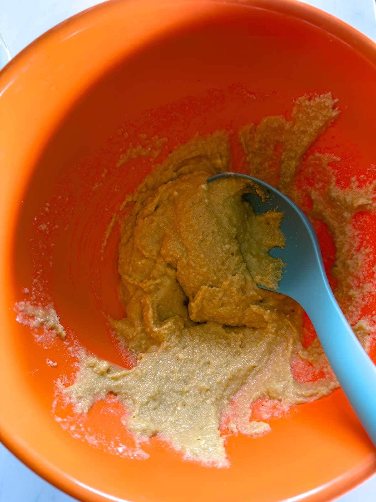 A blue spoon rests in an orange mixing bowl containing a partially mixed, creamy batter or dough.