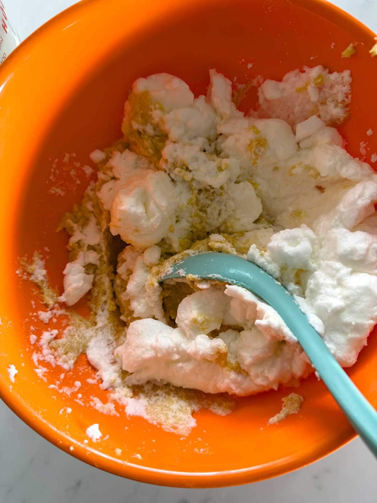 A teal spatula mixes fluffy egg whites or meringue into a brown batter inside a bright orange mixing bowl.
