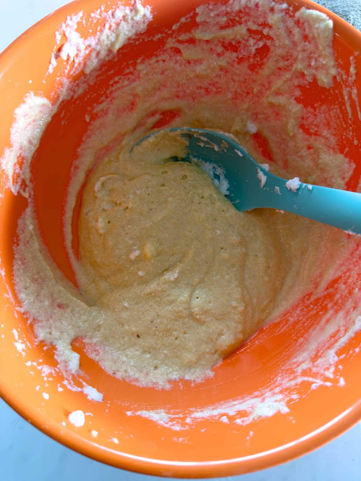 A bright orange mixing bowl containing thick, pale batter being mixed with a blue silicone spatula. Batter is sticking to the sides of the bowl.