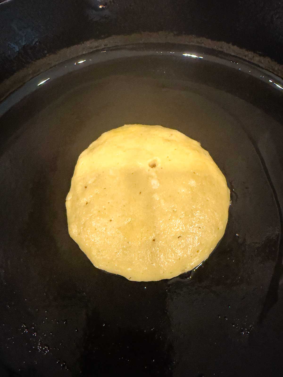 A pancake in a black cast iron pan with oil, beginning to puff up and turn golden brown.