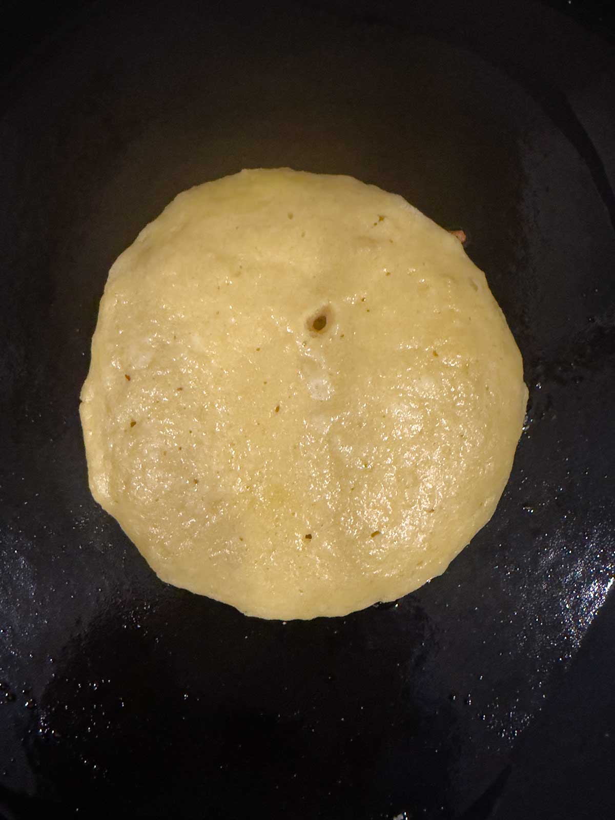 A pancake is cooking on a dark, lightly oiled surface, viewed from above.