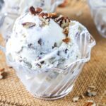 A close-up of a glass bowl filled with vanilla ice cream mixed with chopped pecans, topped with extra pecans, placed on a textured brown surface.