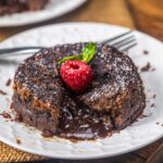A chocolate lava cake on a white plate, with rich molten chocolate oozing from the center. It’s topped with a fresh raspberry, a mint leaf, and powdered sugar. A fork rests beside the dessert.