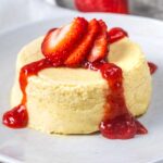 A creamy, round cheesecake topped with sliced fresh strawberries and drizzled with bright red strawberry sauce, served on a white plate.