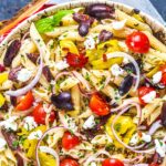 A bowl of keto pasta salad with cherry tomatoes, sliced red onions, black olives, banana peppers, feta cheese, and fresh herbs, arranged on a wooden tray.