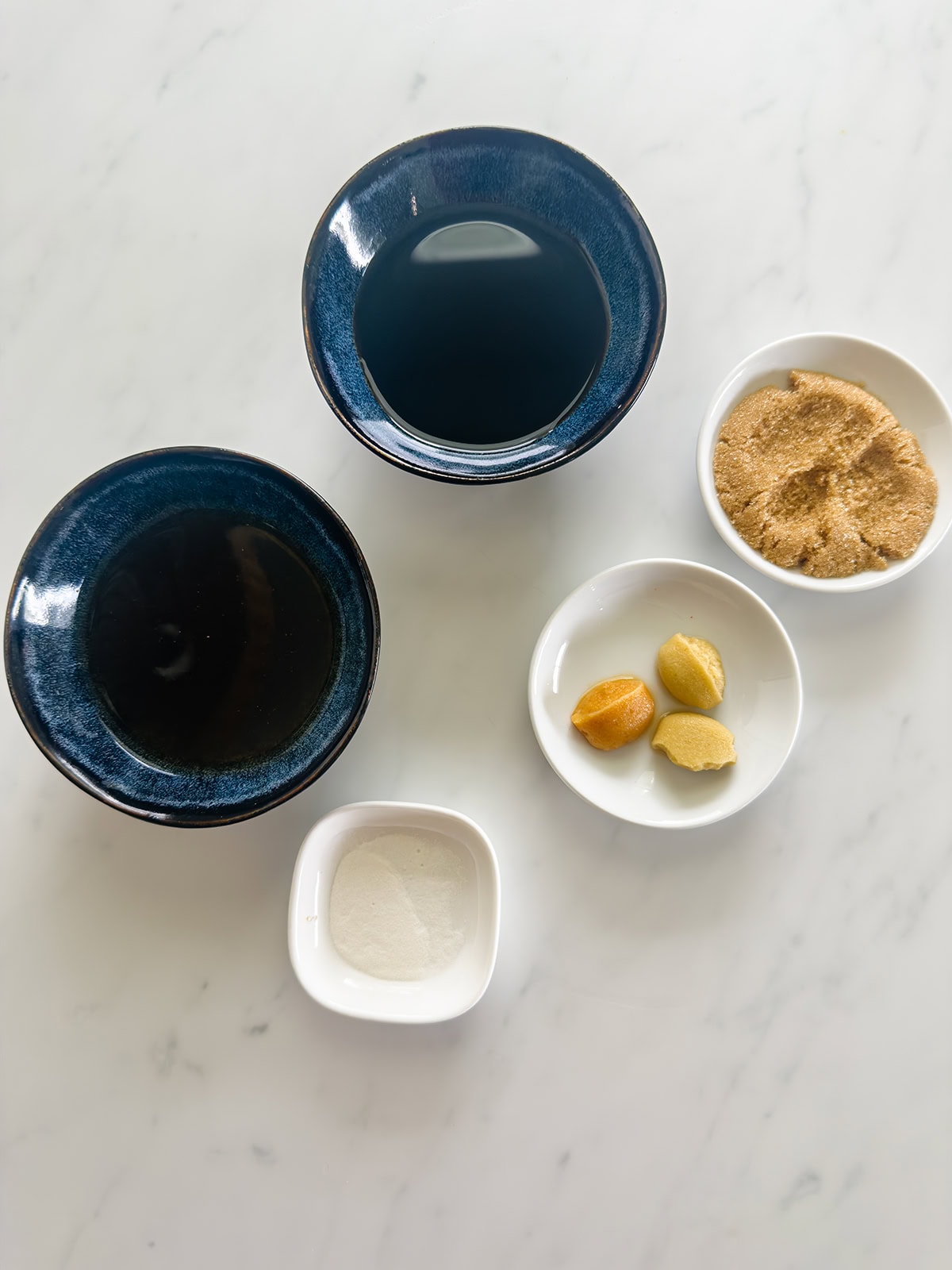 Five small bowls on a white marble surface, each holding key ingredients—coconut aminos, garlic, ginger, and sweetener—perfect for making a flavorful keto chicken stir fry.