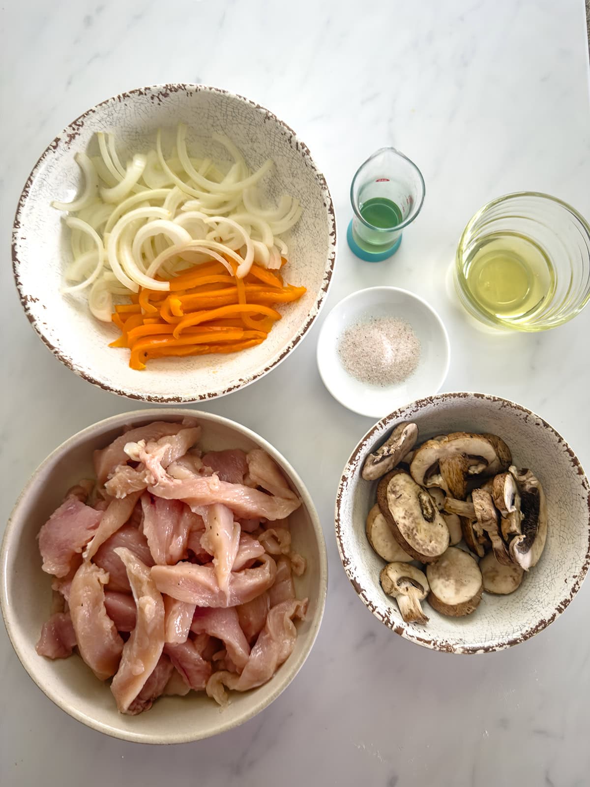 Four bowls on a white surface display ingredients for a keto chicken stir fry, including sliced onions and bell peppers, raw chicken strips, mushrooms, seasoning, and two small glasses of liquid ingredients.