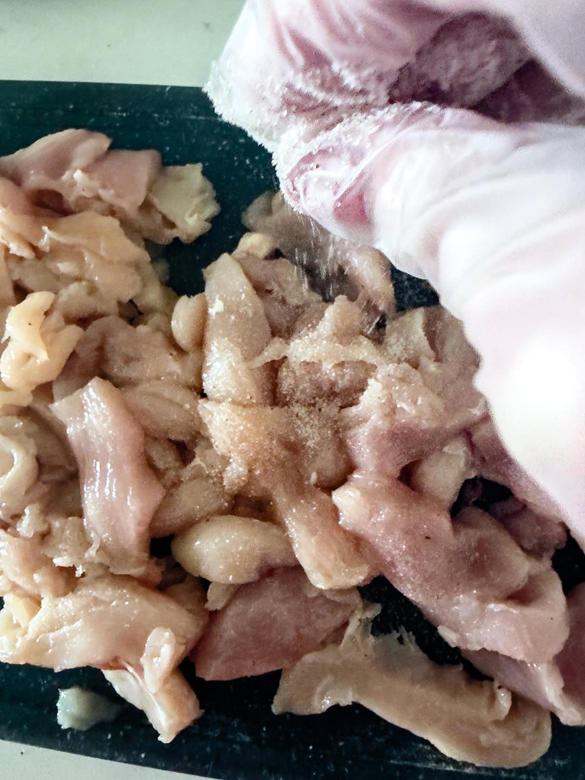 A gloved hand sprinkling seasoning onto raw, sliced chicken pieces on a dark surface, preparing the meat for a flavorful keto chicken stir fry.