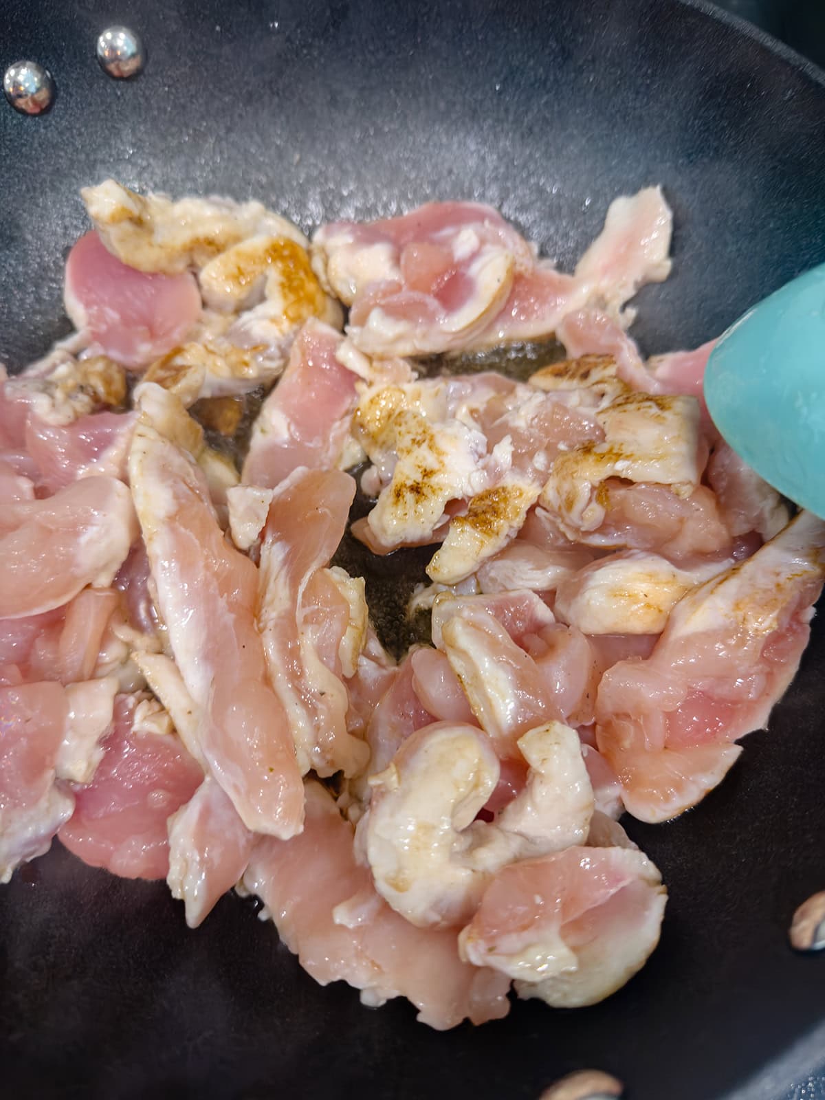 Raw chicken slices for a keto chicken stir fry are being cooked in a black frying pan, with some pieces turning white from the heat while others remain pink. A teal utensil is partially visible on the right side of the image.