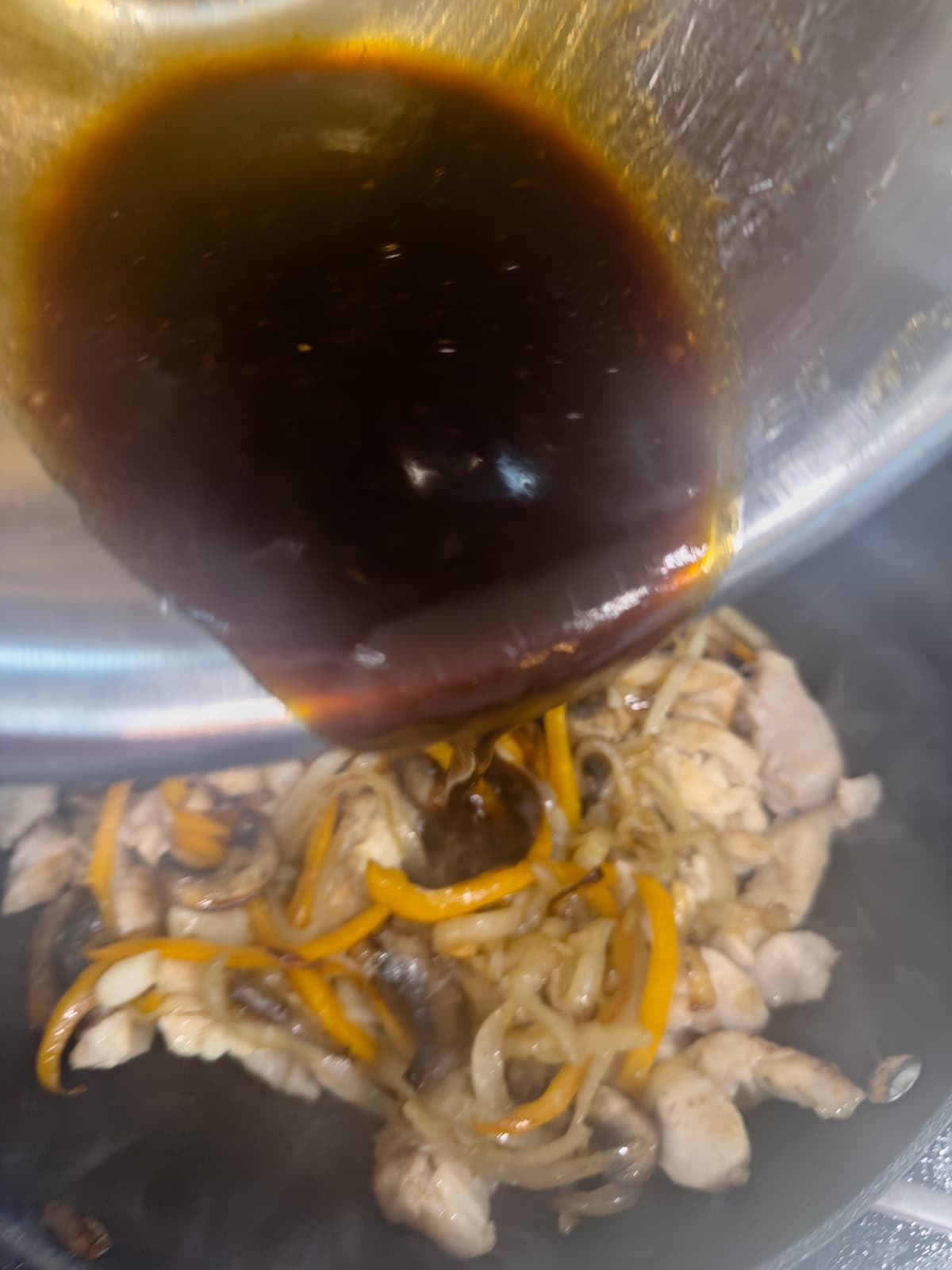 A dark sauce is being poured from a bowl onto a keto chicken stir fry mixture of sliced chicken, mushrooms, onions, and yellow bell peppers in a pan.
