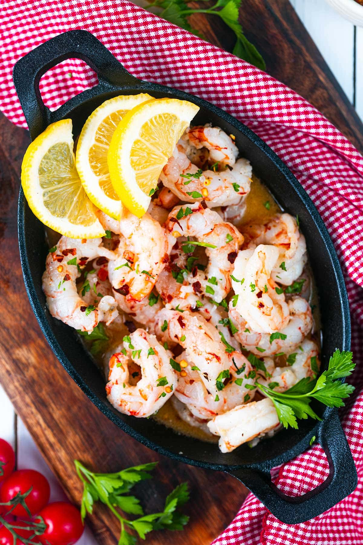 A black dish filled with cooked shrimp garnished with chopped herbs, red pepper flakes, and three lemon slices, set on a wooden board with a red and white checkered cloth. Fresh parsley and tomatoes are nearby.