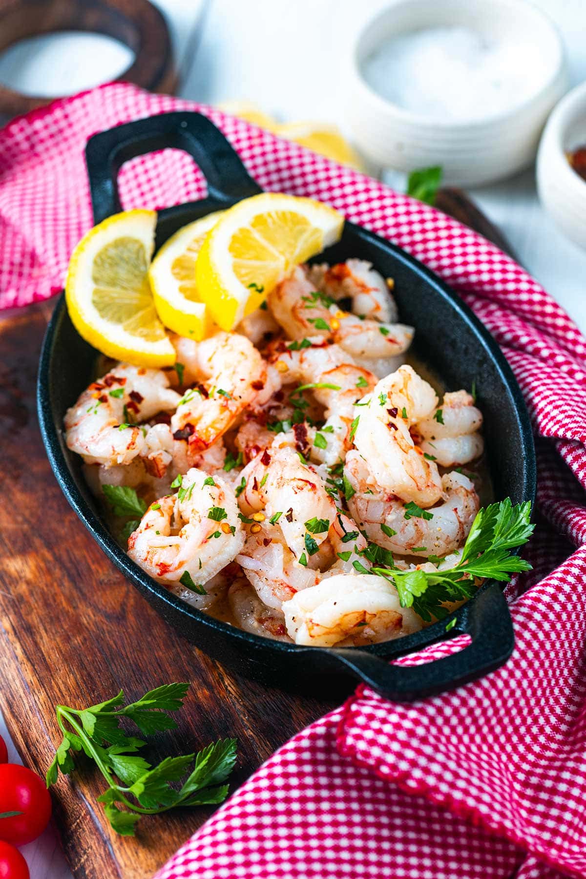 A black skillet filled with cooked shrimp, garnished with chopped parsley and three lemon slices, sits on a wooden board next to a red checkered cloth. Fresh parsley and small bowls are in the background.