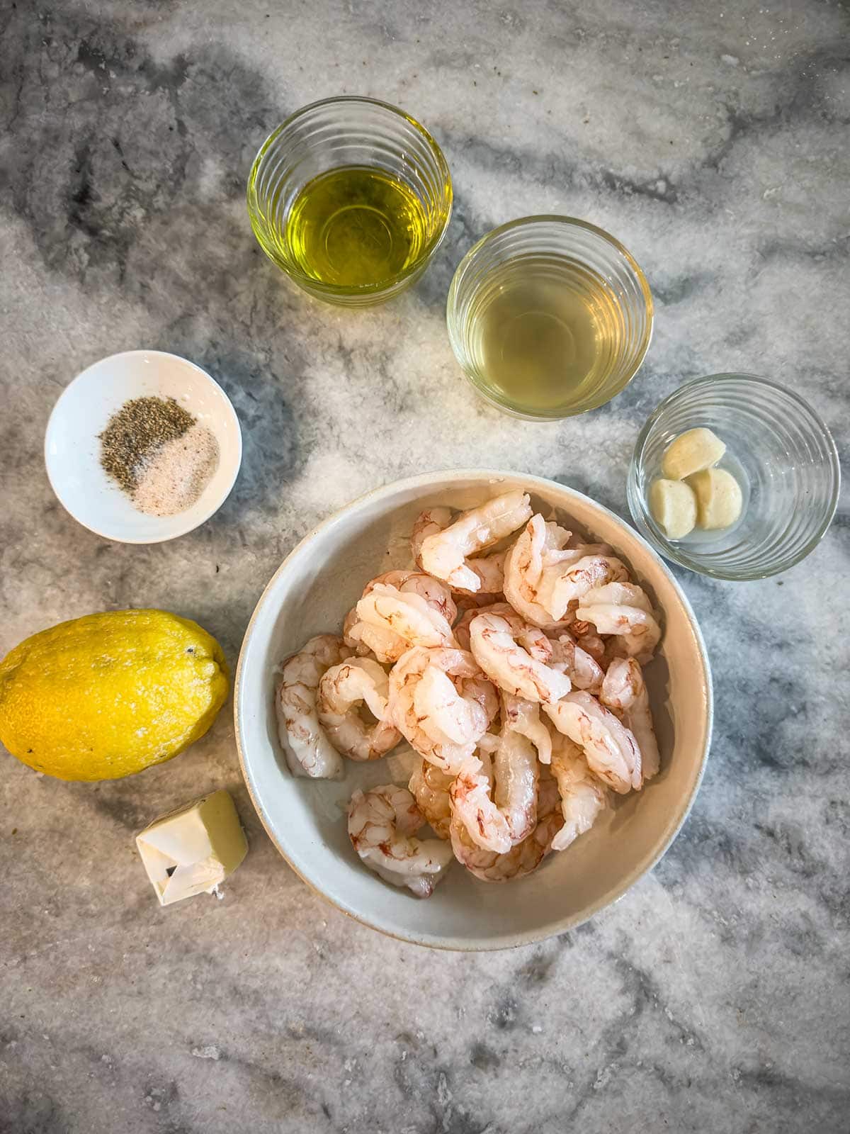 A marble countertop with raw shrimp in a bowl, a lemon, two garlic cloves in a small dish, a cube of butter, a small bowl of salt and pepper, and three glass cups containing white wine, olive oil, and water.