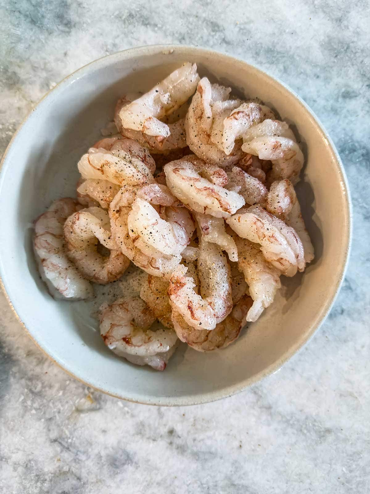 A white bowl filled with uncooked, peeled shrimp seasoned with salt and pepper, placed on a light-colored marble surface.