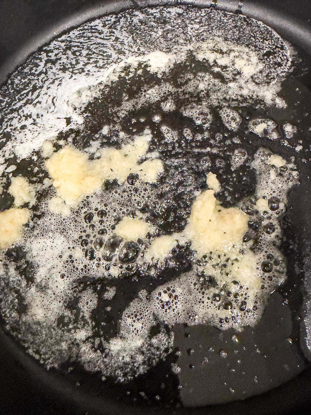 Garlic sautéing in melted butter in a black frying pan, with bubbles forming around the garlic and butter mixture.
