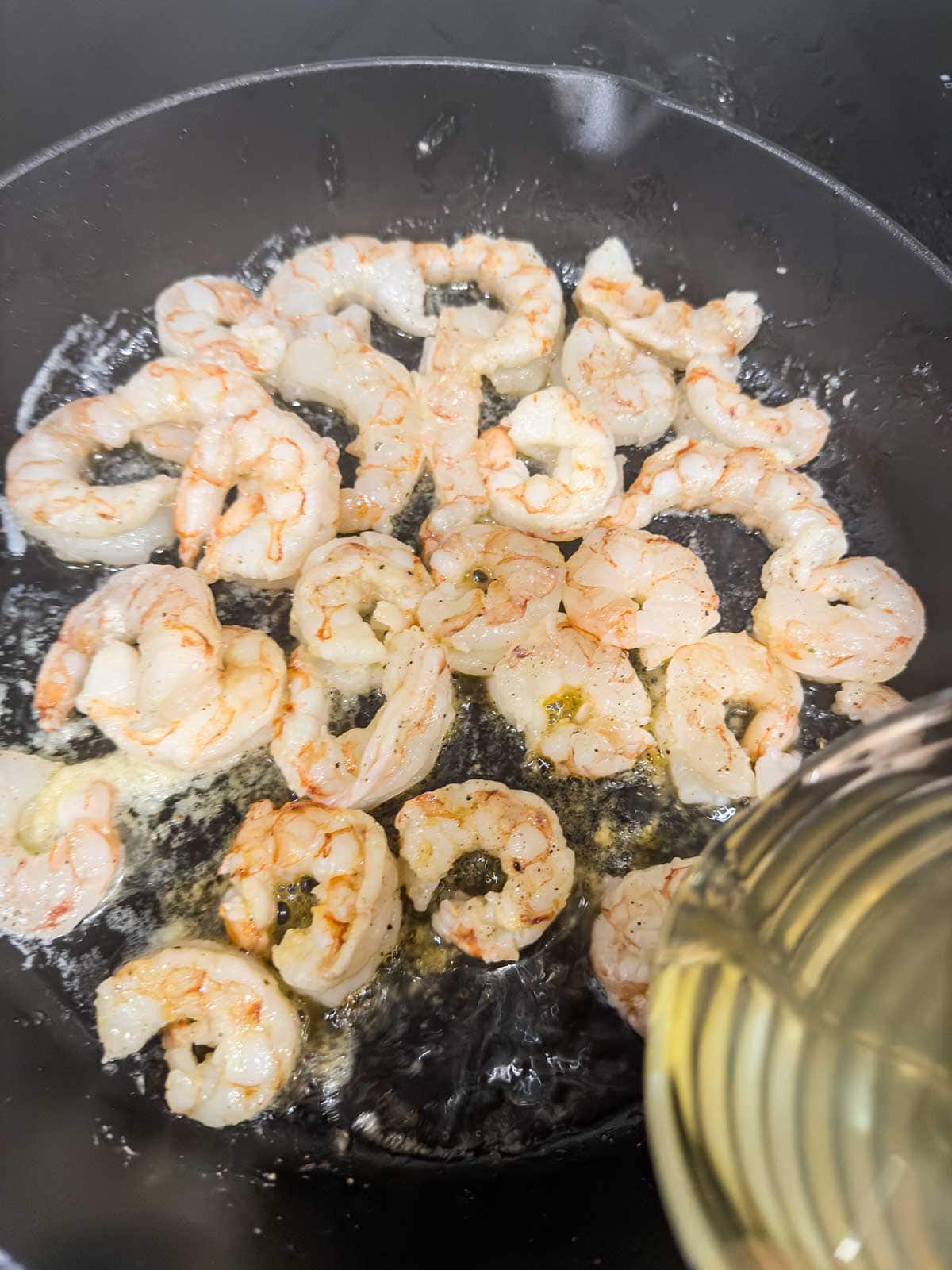 Shrimp being sautéed in a sizzling pan with oil, and a stream of liquid, possibly wine or broth, being poured in from the side.