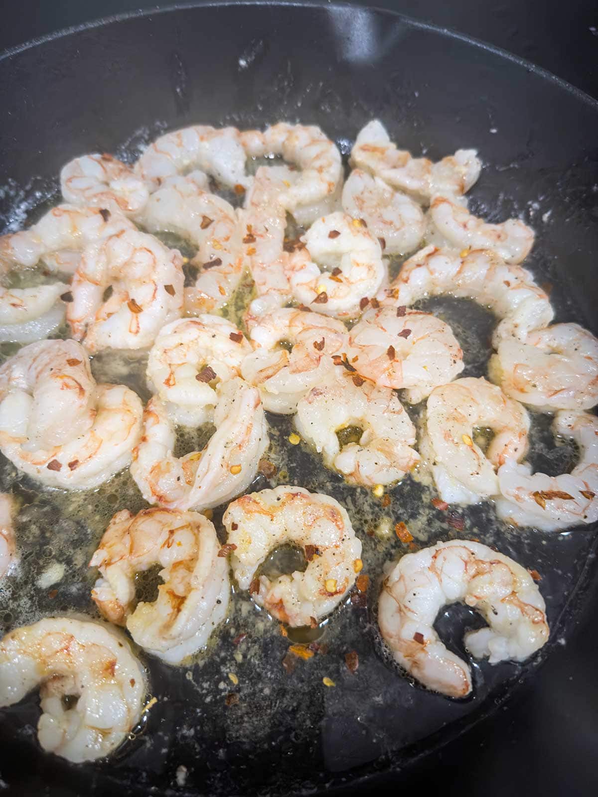 Shrimp cooking in a black skillet with melted butter and minced garlic, sprinkled with red pepper flakes.