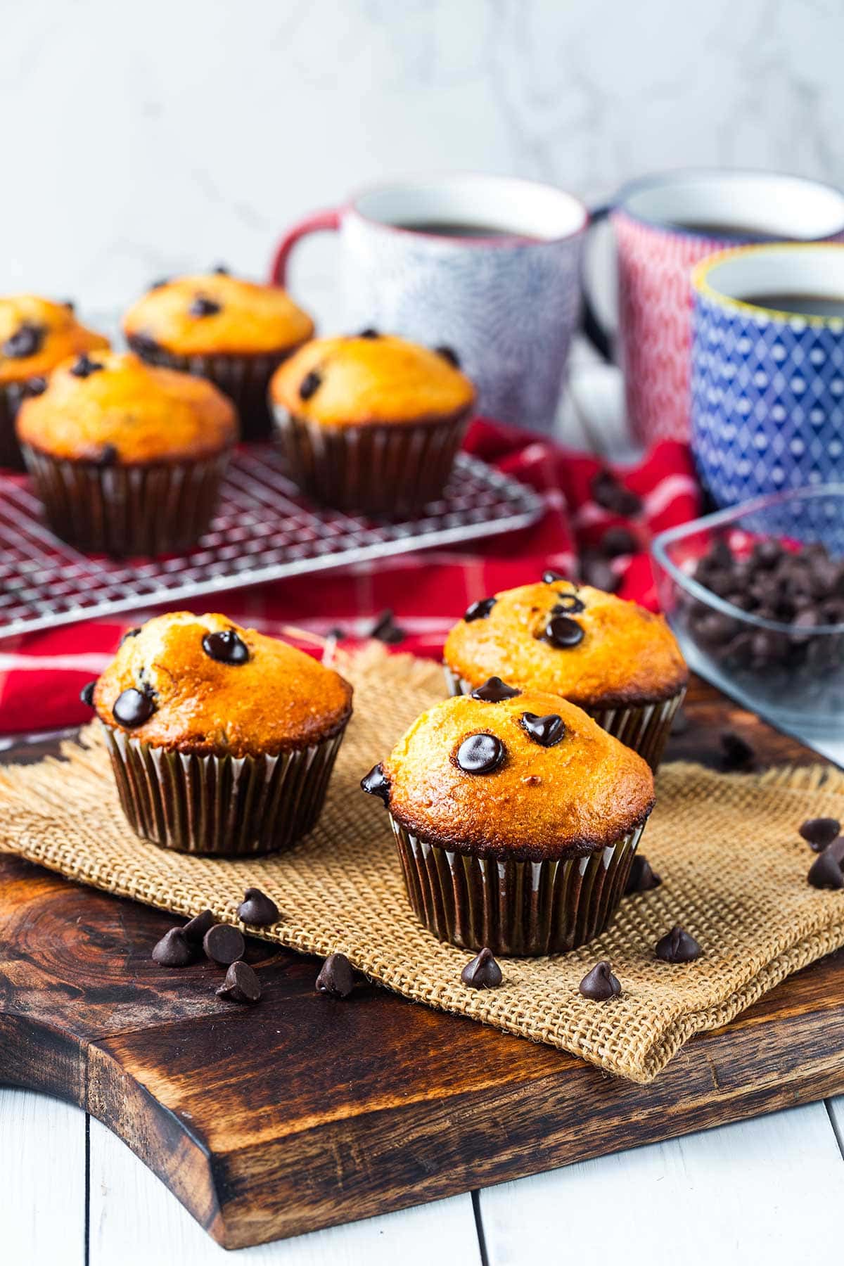 Three chocolate chip muffins sit on a wooden board, with more muffins cooling on a rack in the background. Coffee mugs, a red cloth, and a bowl of chocolate chips are also visible.
