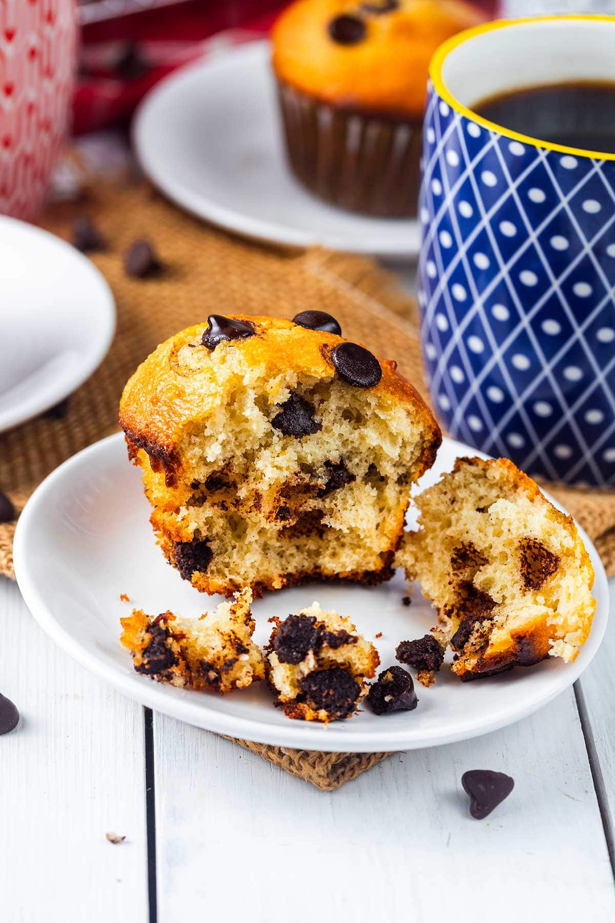 A chocolate chip muffin is split open on a white plate with crumbs and chocolate chips around it. A blue patterned mug of coffee and another muffin are in the background.