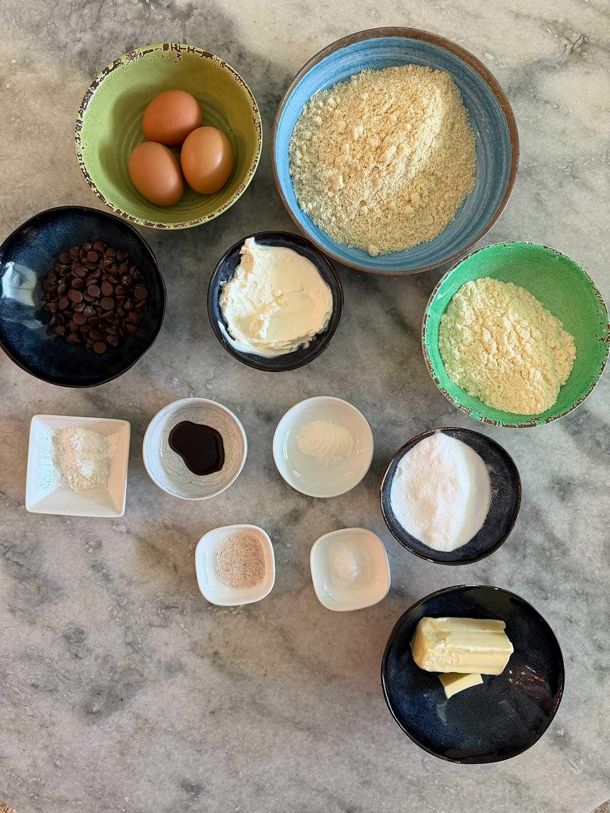 A marble surface with bowls containing eggs, chocolate chips, yogurt, butter, almond flour, protein powder, sugar, vanilla, baking powder, and other baking ingredients arranged neatly.