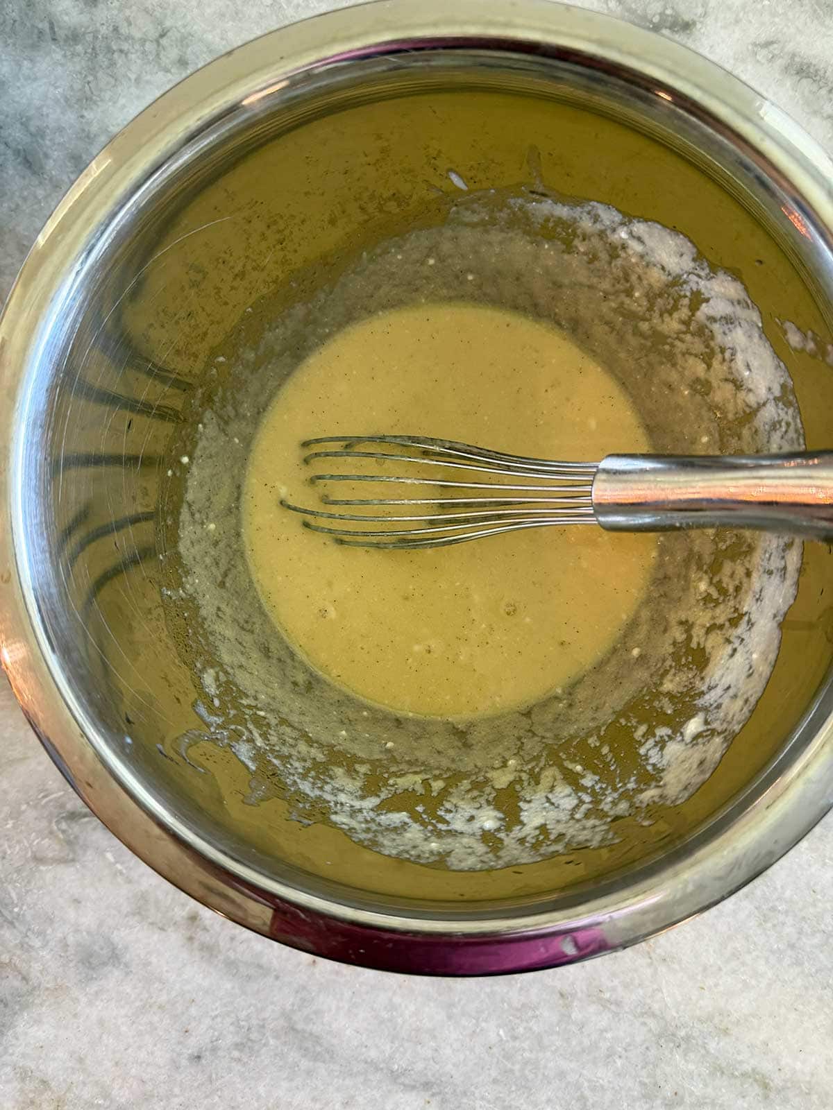 A metal whisk rests in a stainless steel bowl containing a light yellow batter, partially mixed, sitting on a marble countertop.