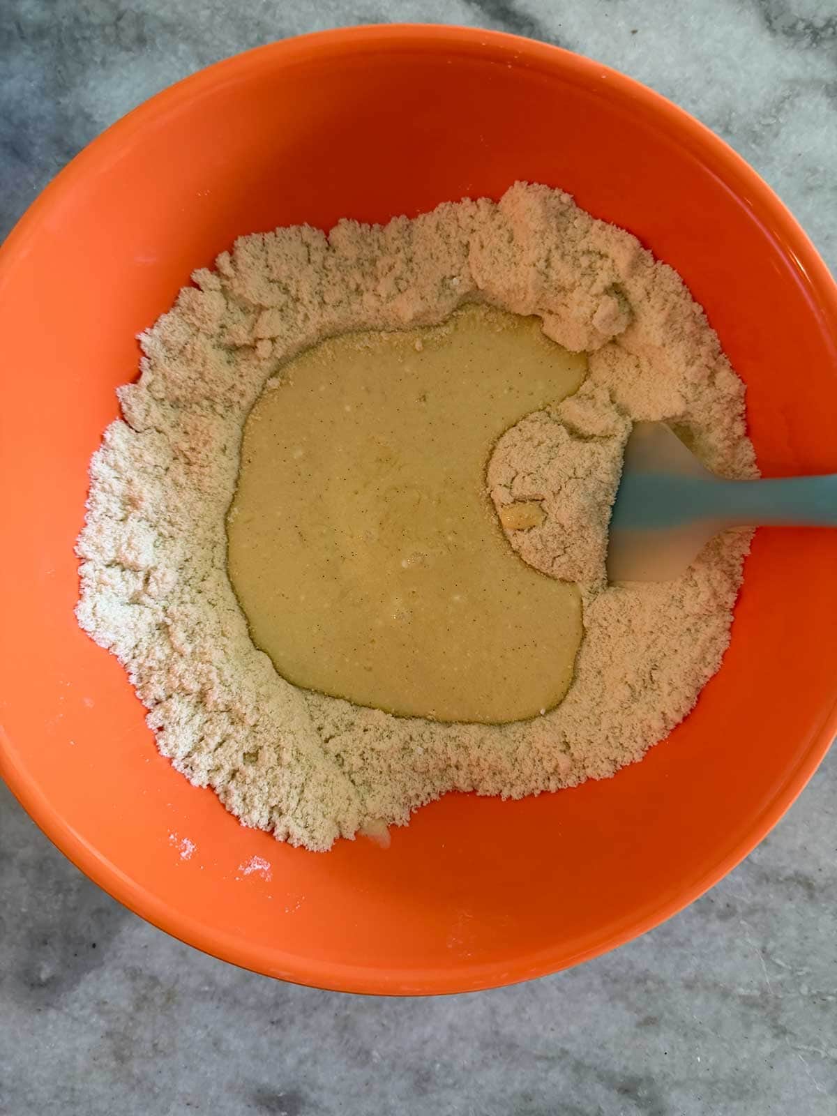 An orange bowl with dry ingredients and a well in the center filled with liquid ingredients, being mixed with a blue spatula on a gray countertop.