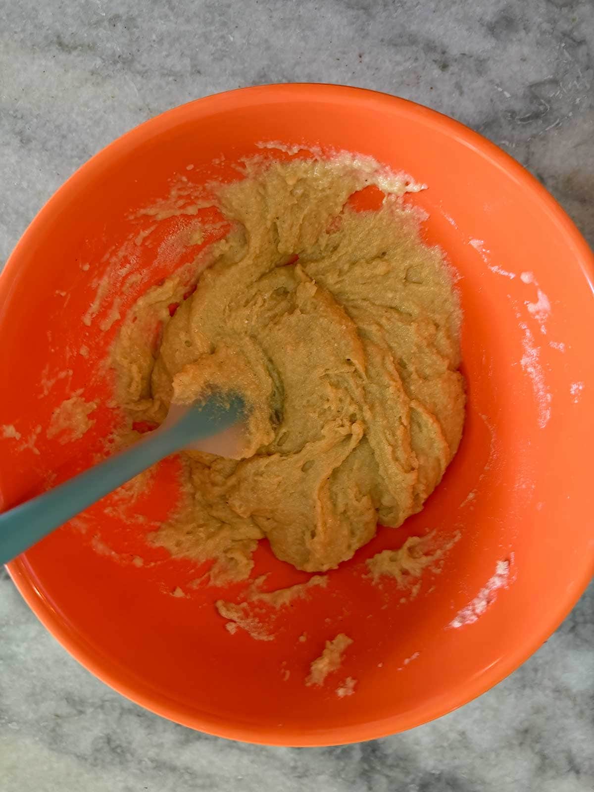 Orange bowl with a thick, beige batter being mixed using a blue spatula, placed on a gray marble countertop. The batter appears partially mixed, with some flour visible on the sides of the bowl.