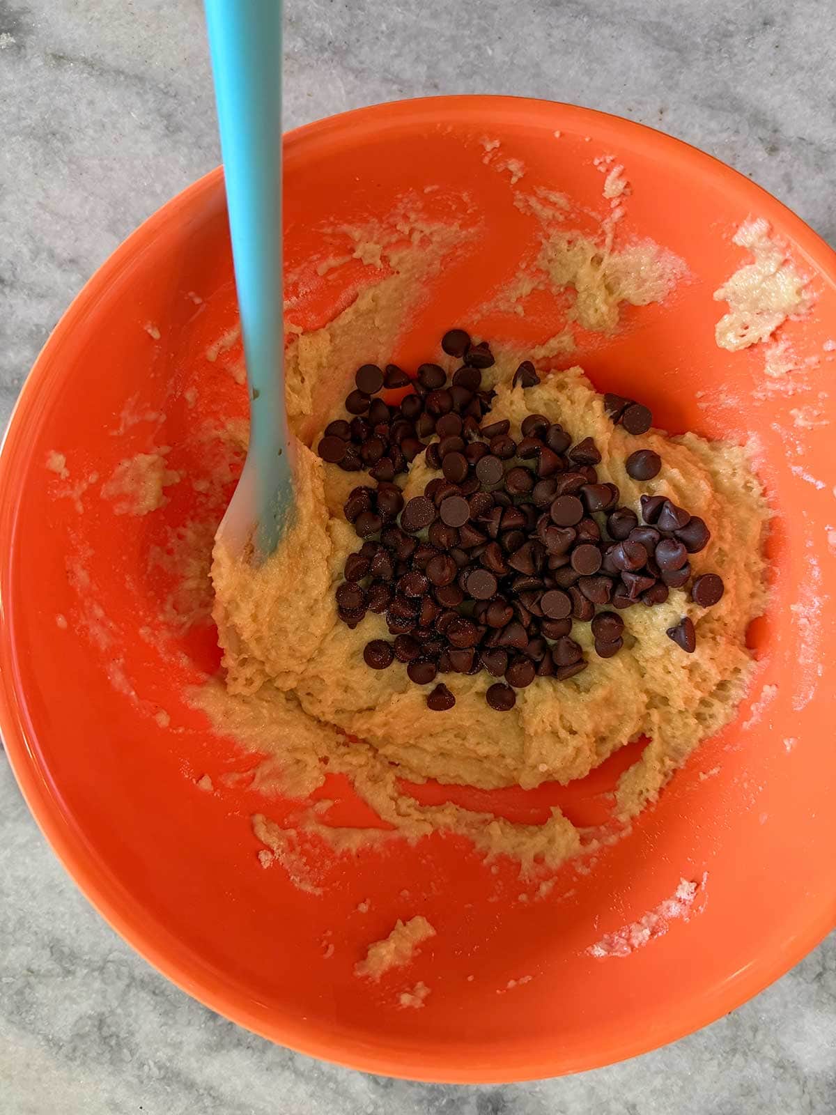 An orange bowl with cookie dough and chocolate chips, being mixed with a blue spatula, sits on a gray countertop.