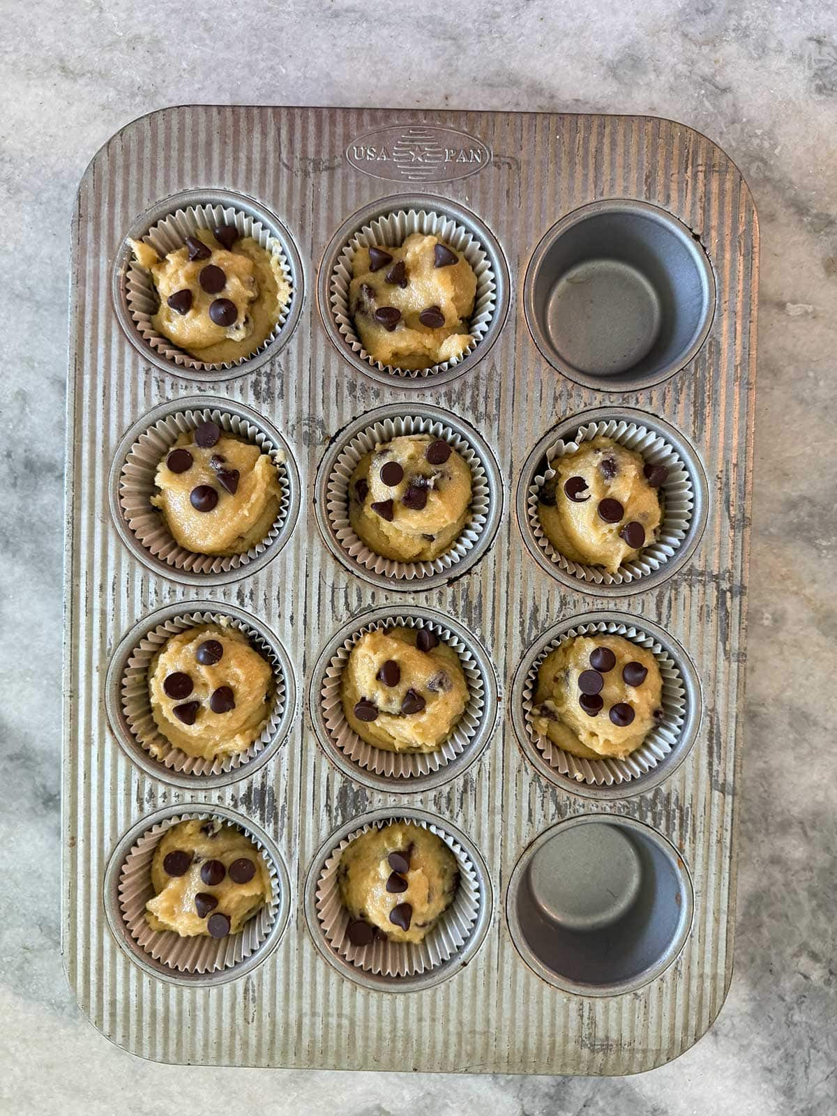 A muffin tin on a marble surface holds nine unbaked batter-filled cups topped with chocolate chips; three spaces are empty.