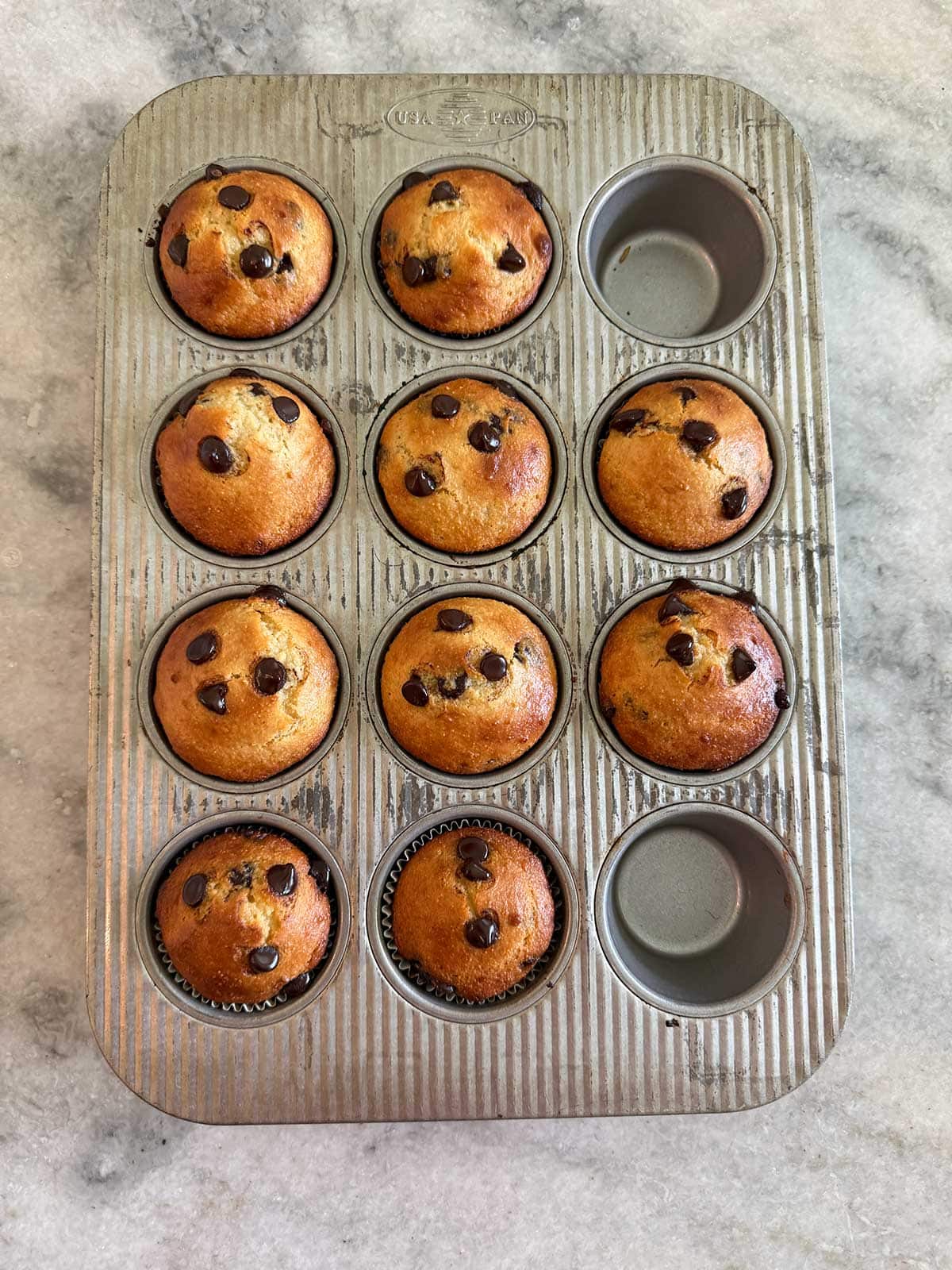 A metal muffin tray with ten baked chocolate chip muffins and two empty slots, placed on a light gray marble countertop.