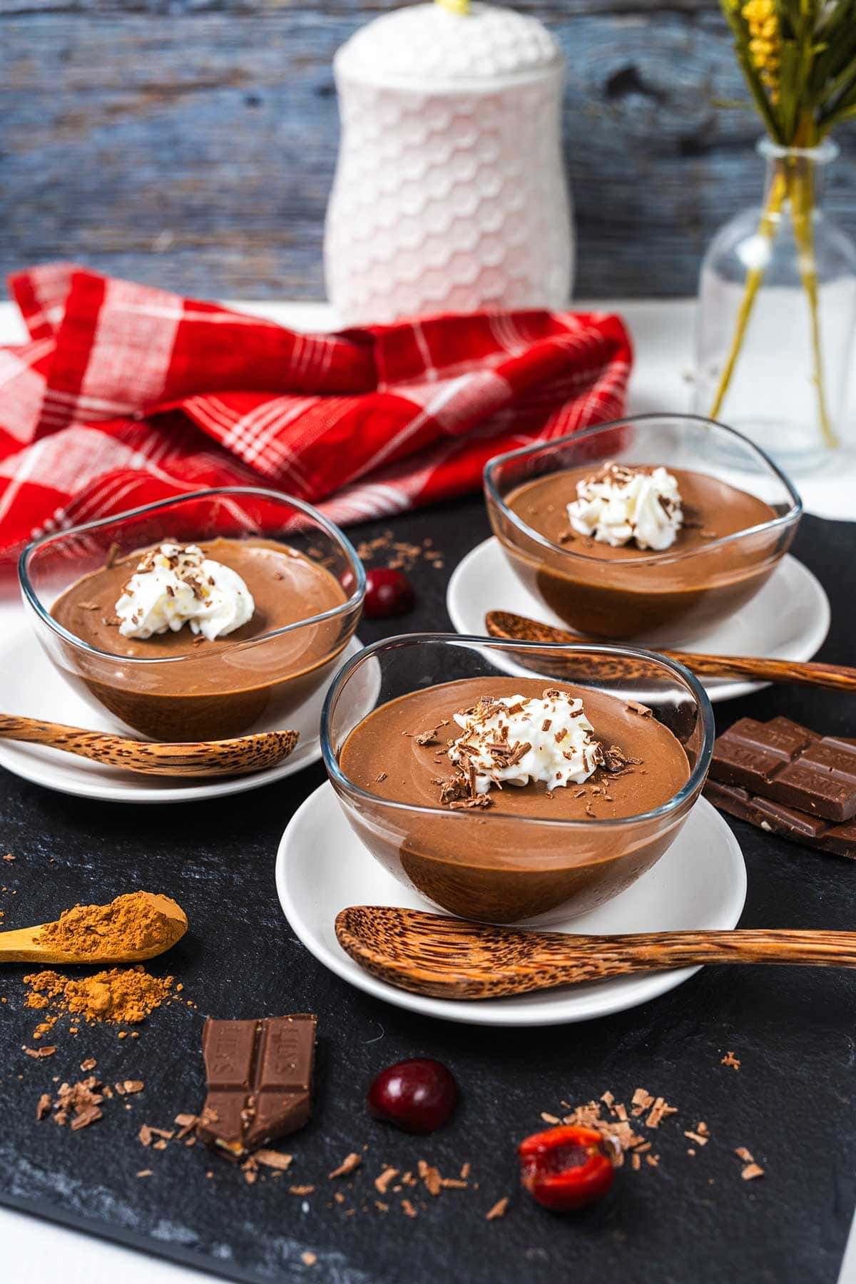 Three glass bowls of chocolate mousse topped with whipped cream and chocolate shavings, each on a white plate with a wooden spoon, surrounded by chocolate pieces, cocoa powder, cherries, and a red checkered cloth.