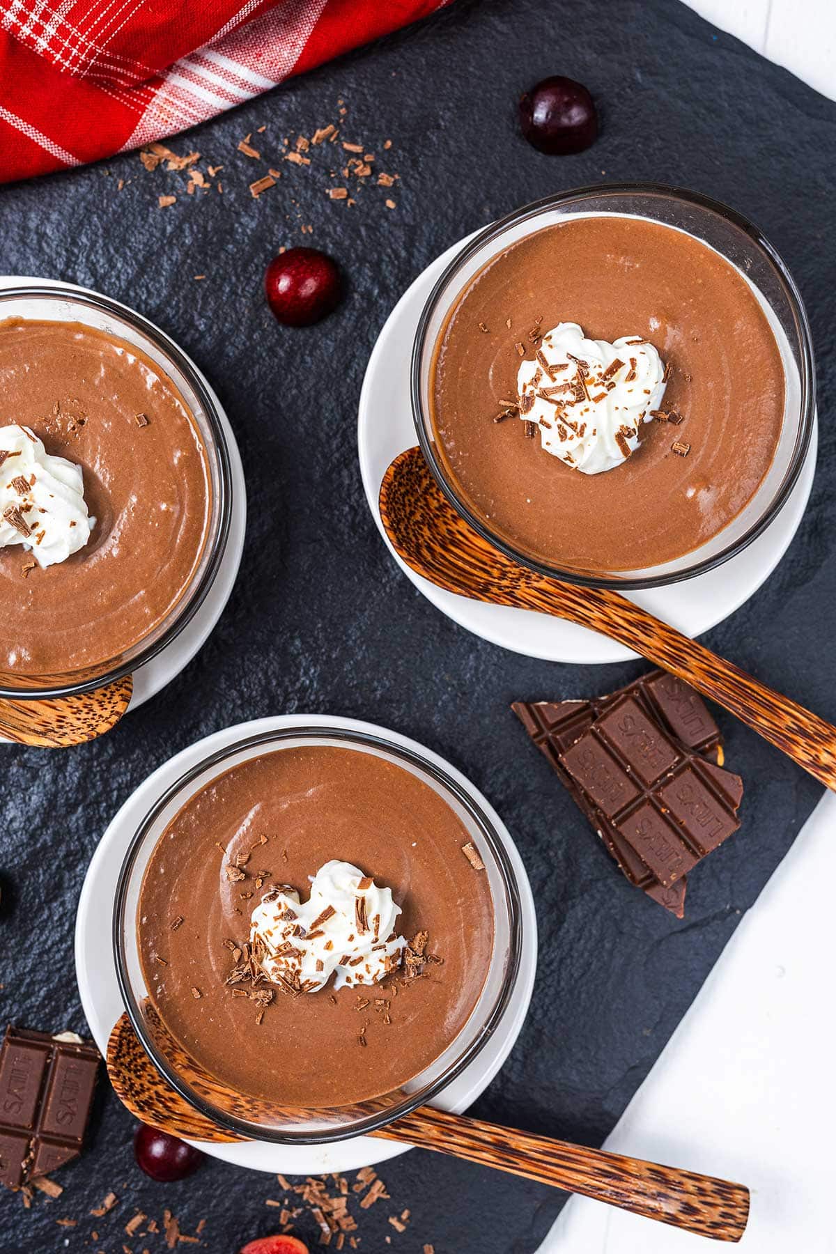 Three bowls of chocolate mousse topped with whipped cream and chocolate shavings, each with a wooden spoon, sit on white saucers. Pieces of chocolate and cherries are scattered on a dark slate surface nearby.