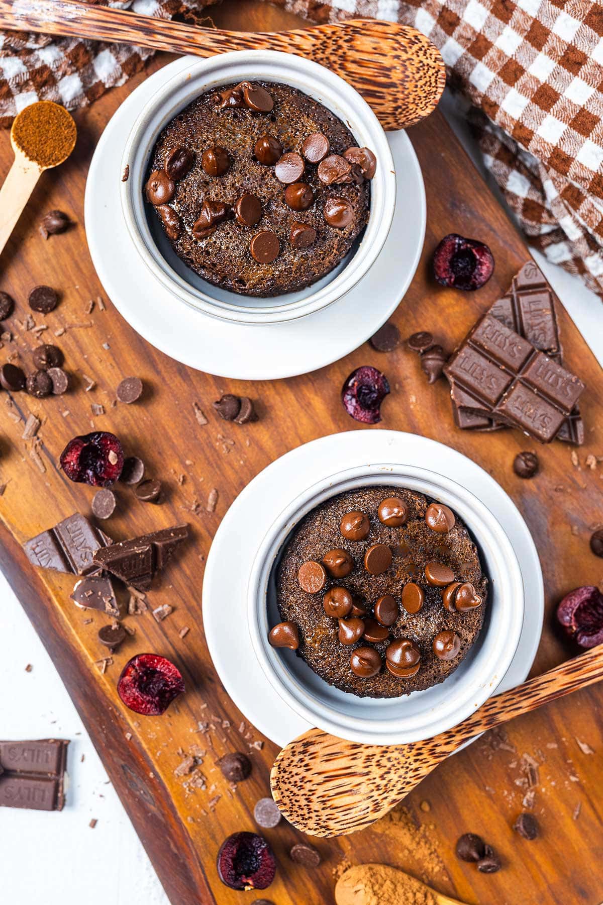 Two bowls of chocolate mug cake topped with chocolate chips sit on white saucers, surrounded by chocolate pieces, dried cherries, a brown checked napkin, and wooden spoons on a wooden board.