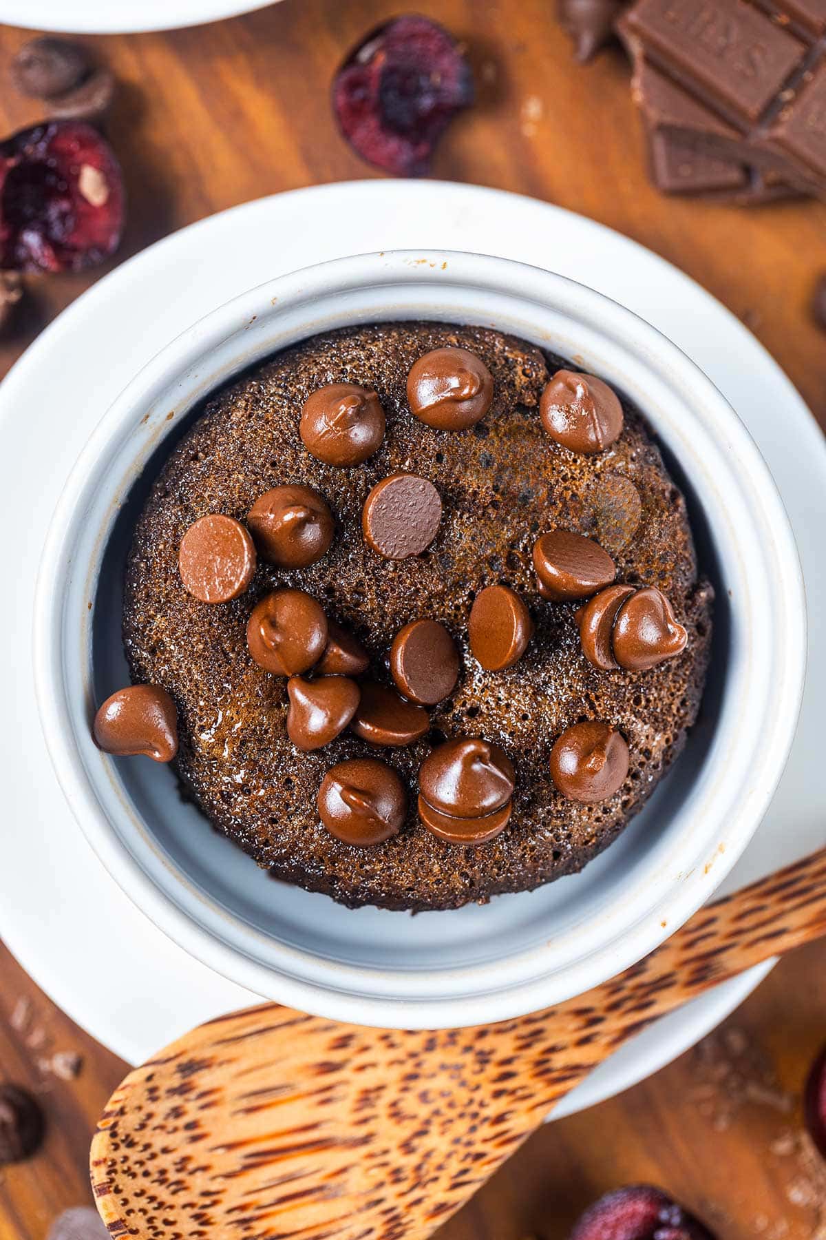 A chocolate mug cake topped with melted chocolate chips sits in a white ramekin on a saucer, with a wooden spoon beside it and chocolate pieces and cherries scattered nearby.