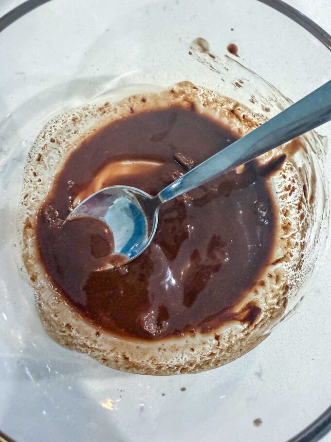 A metal spoon rests in a glass bowl containing a mixture reminiscent of melted chocolate and cream for a delicious Keto Mug Cake, partially stirred, on a light-colored surface.