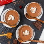 Three glass bowls of chocolate mousse topped with whipped cream and chocolate shavings, each with a wooden spoon. Nearby are chocolate pieces, cocoa powder, a cherry, and a red napkin on a slate serving tray.