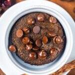A close-up of a chocolate mug cake topped with melted chocolate chips, served in a white ramekin on a white plate.