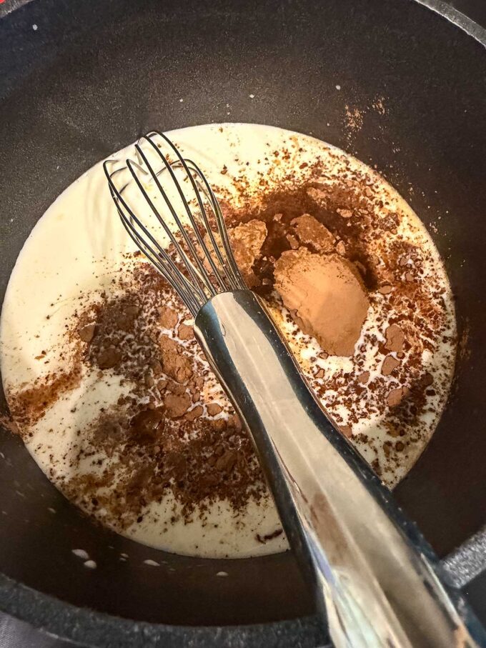 A metal whisk rests in a pot containing cream and cocoa powder, ready to be mixed for a delicious chocolate protein pudding recipe.