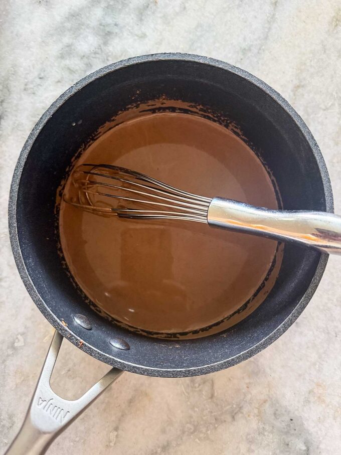 A stainless steel whisk rests in a black saucepan filled with rich chocolate protein pudding, sitting on a light-colored countertop.