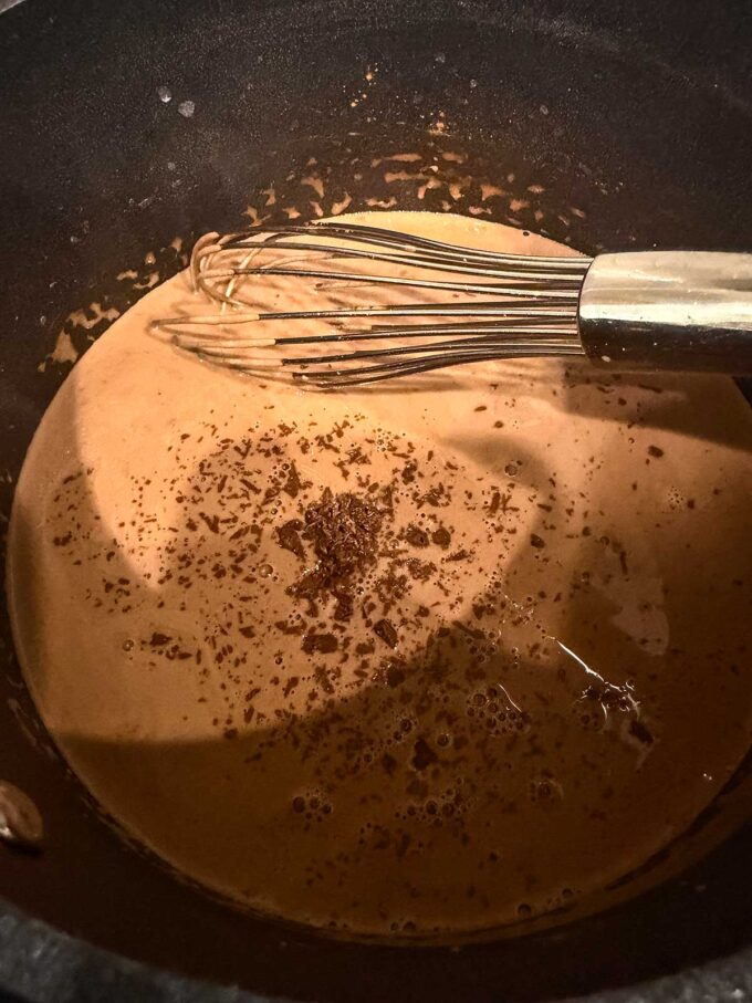 A metal whisk rests in a pot of creamy brown liquid with small chocolate shavings sprinkled on top, suggesting a delicious chocolate protein pudding mixture being prepared.