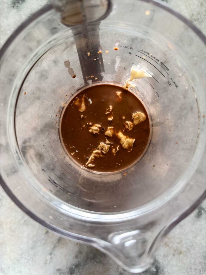 Overhead view of a clear measuring cup containing a small amount of brown liquid—reminiscent of chocolate protein pudding—with dollops of what appears to be mustard or another condiment, sitting on a gray countertop.