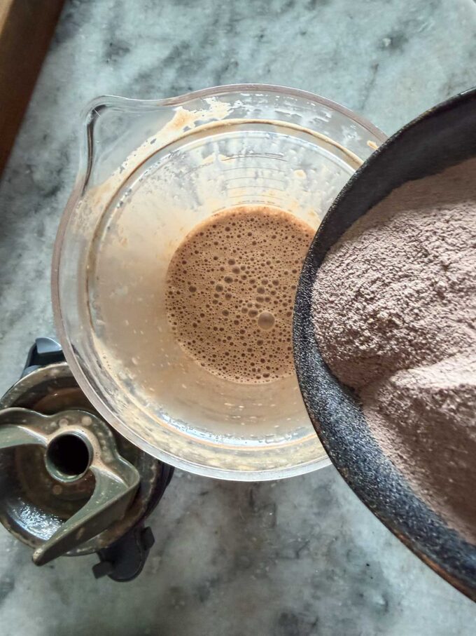 A bowl of chocolate powder is being poured into a blender pitcher containing a frothy liquid, ready to be transformed into rich chocolate protein pudding, with the blender blade attachment placed nearby on a marble countertop.