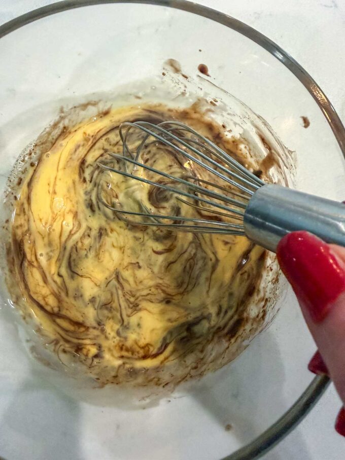 A hand with red nails holds a whisk, mixing a brown and yellow batter for a Keto Dessert in a glass bowl on a light surface.